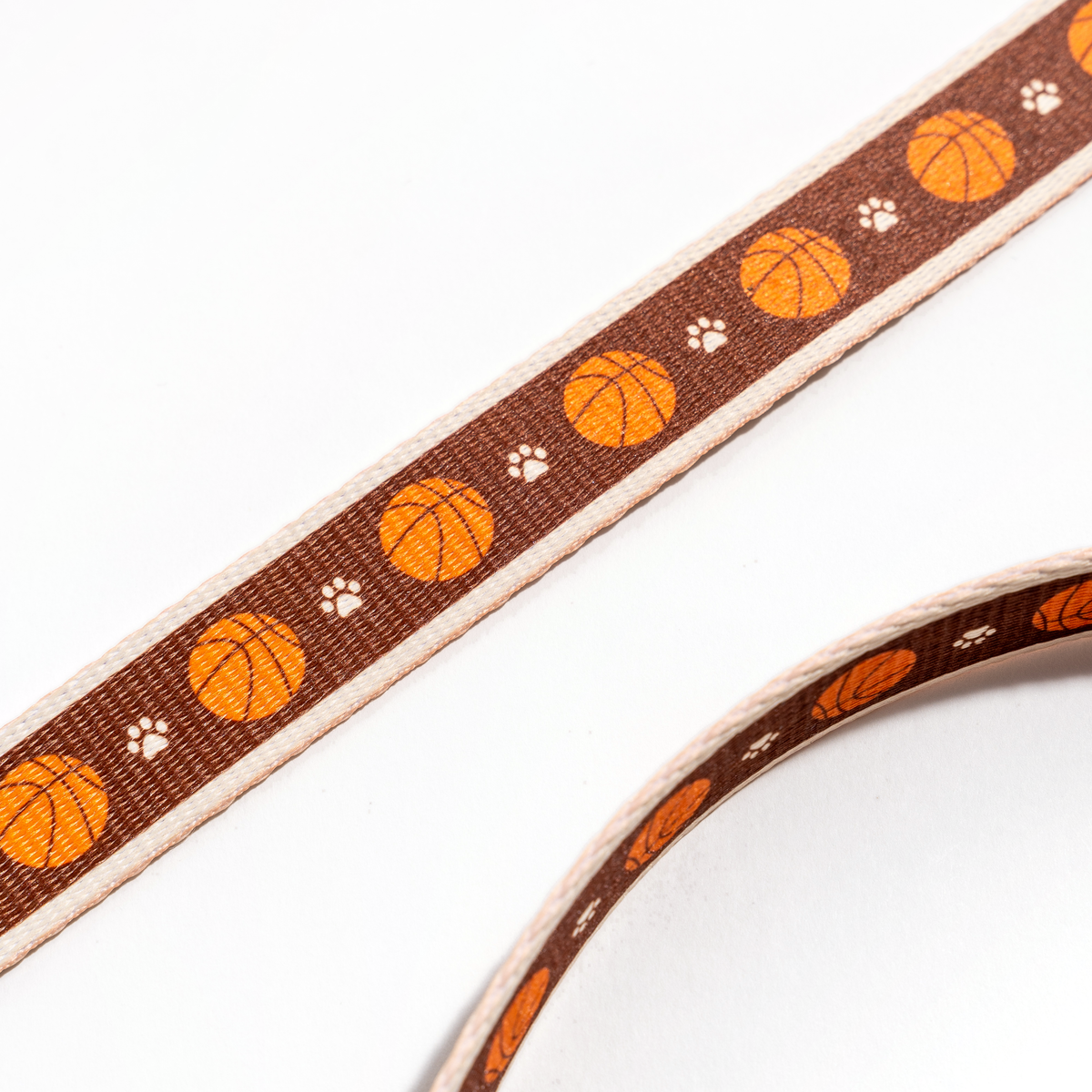 Close-up of the Floofball basketball dog leash featuring repeating orange basketballs and paw prints on brown webbing.