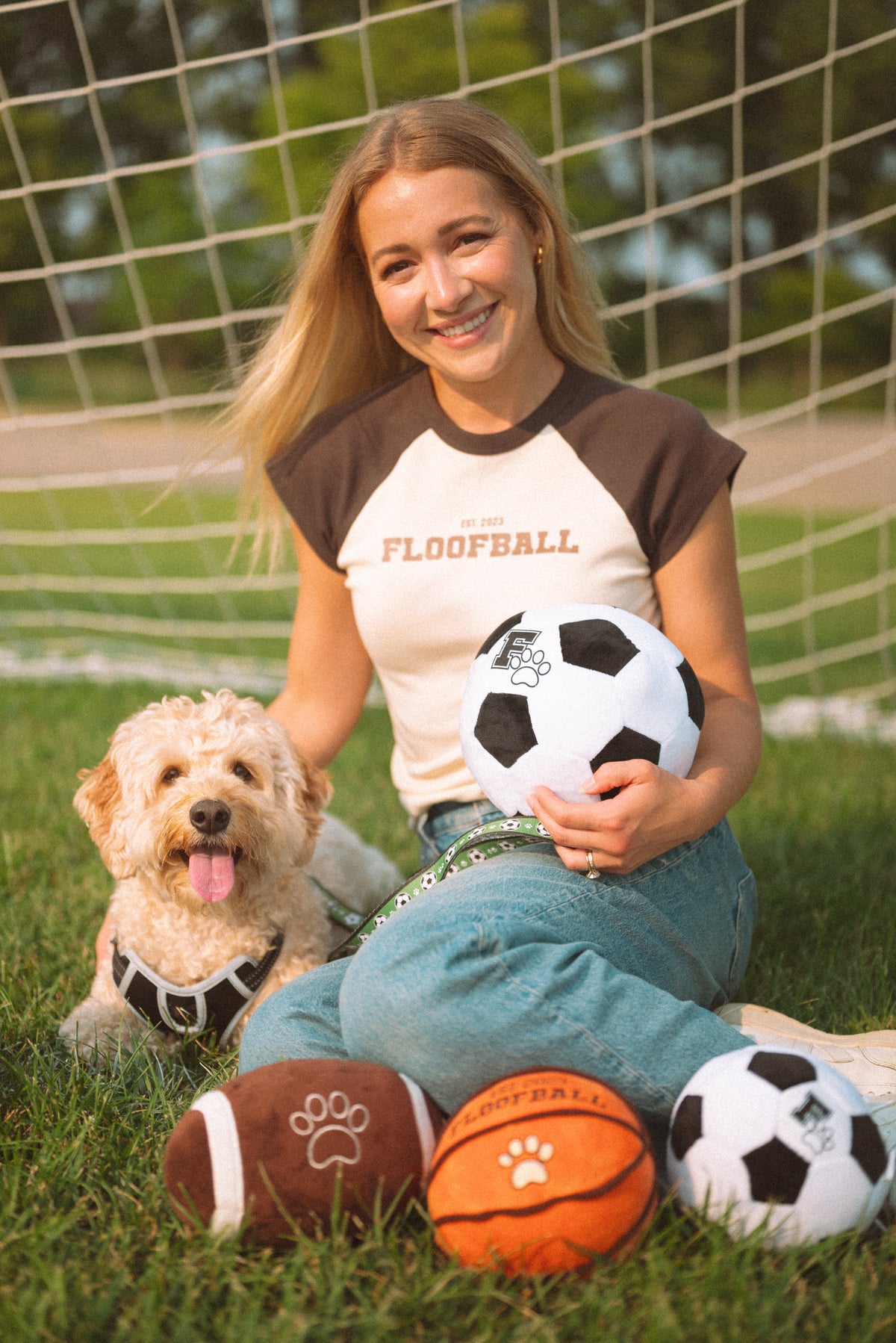 Floofball founder sitting with a dog and sports-inspired dog toys, including plush soccer balls, a basketball, football, and soccer leash on a soccer field.